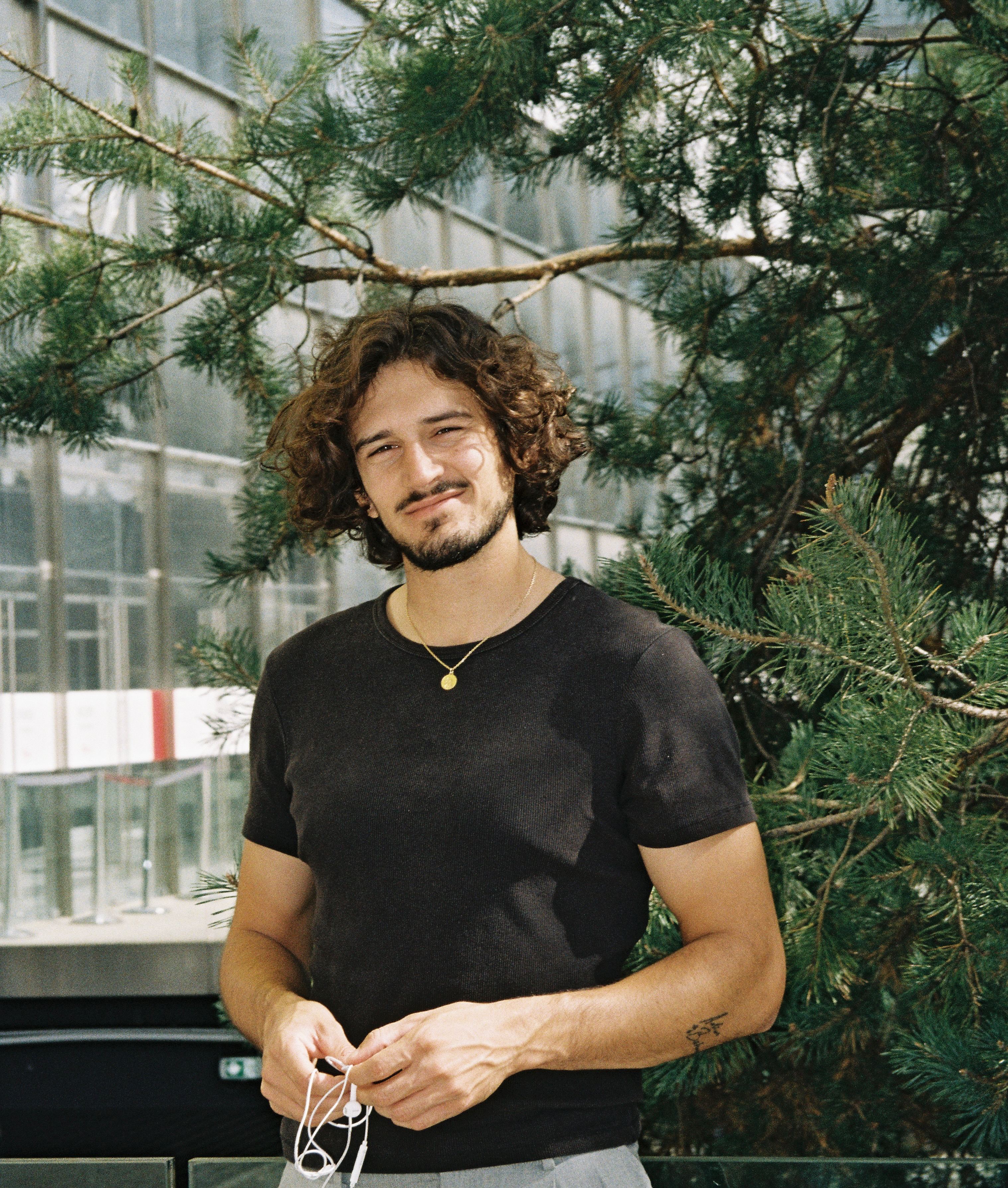Un jeune homme aux cheveux bouclés, t-shirt noir, debout sous les branches d’un pin