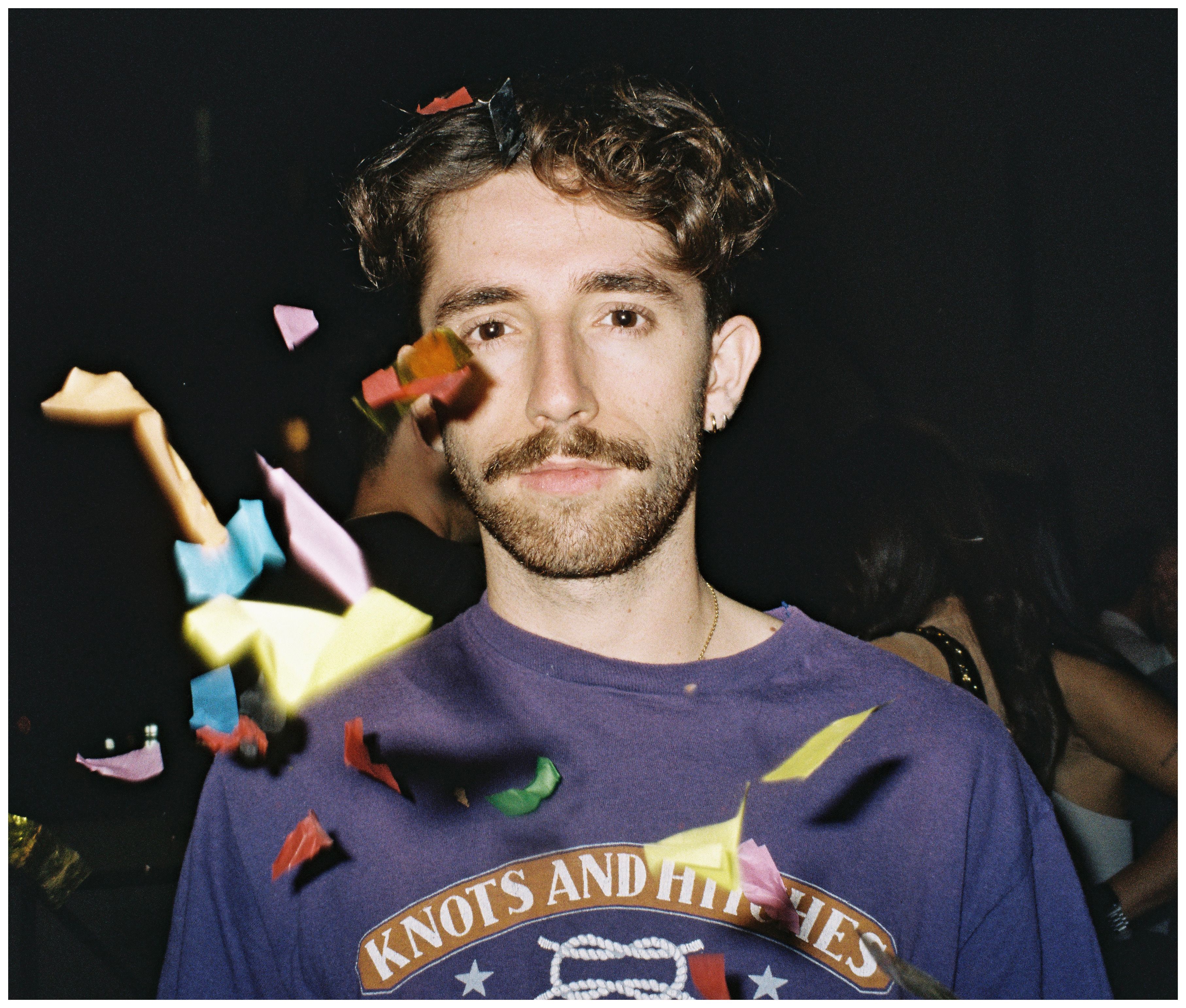Un jeune homme moustachu sous une pluie de confettis, t-shirt violet