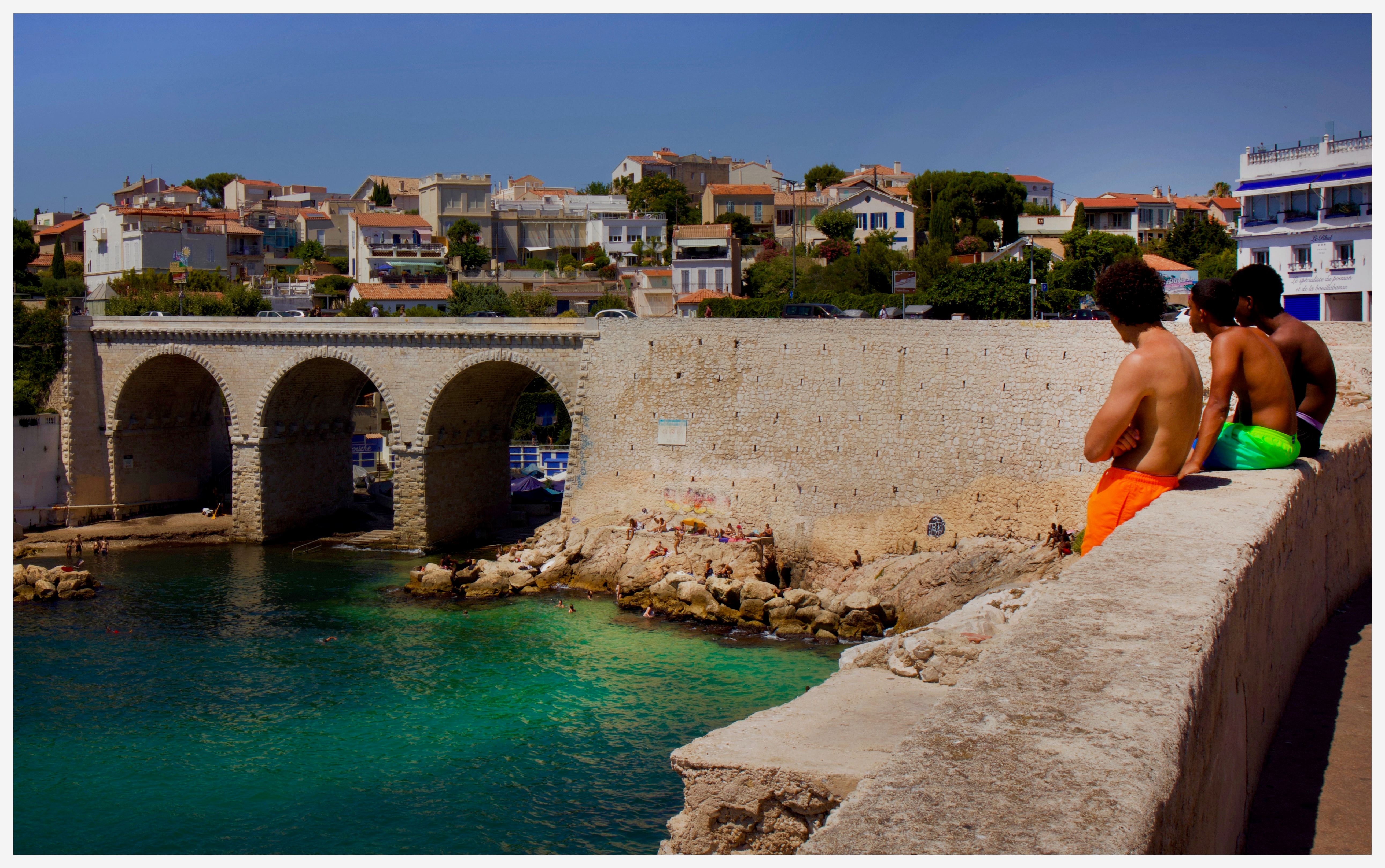 Trois garçons assis sur un muret au-dessus d’une crique, face à un pont à arches