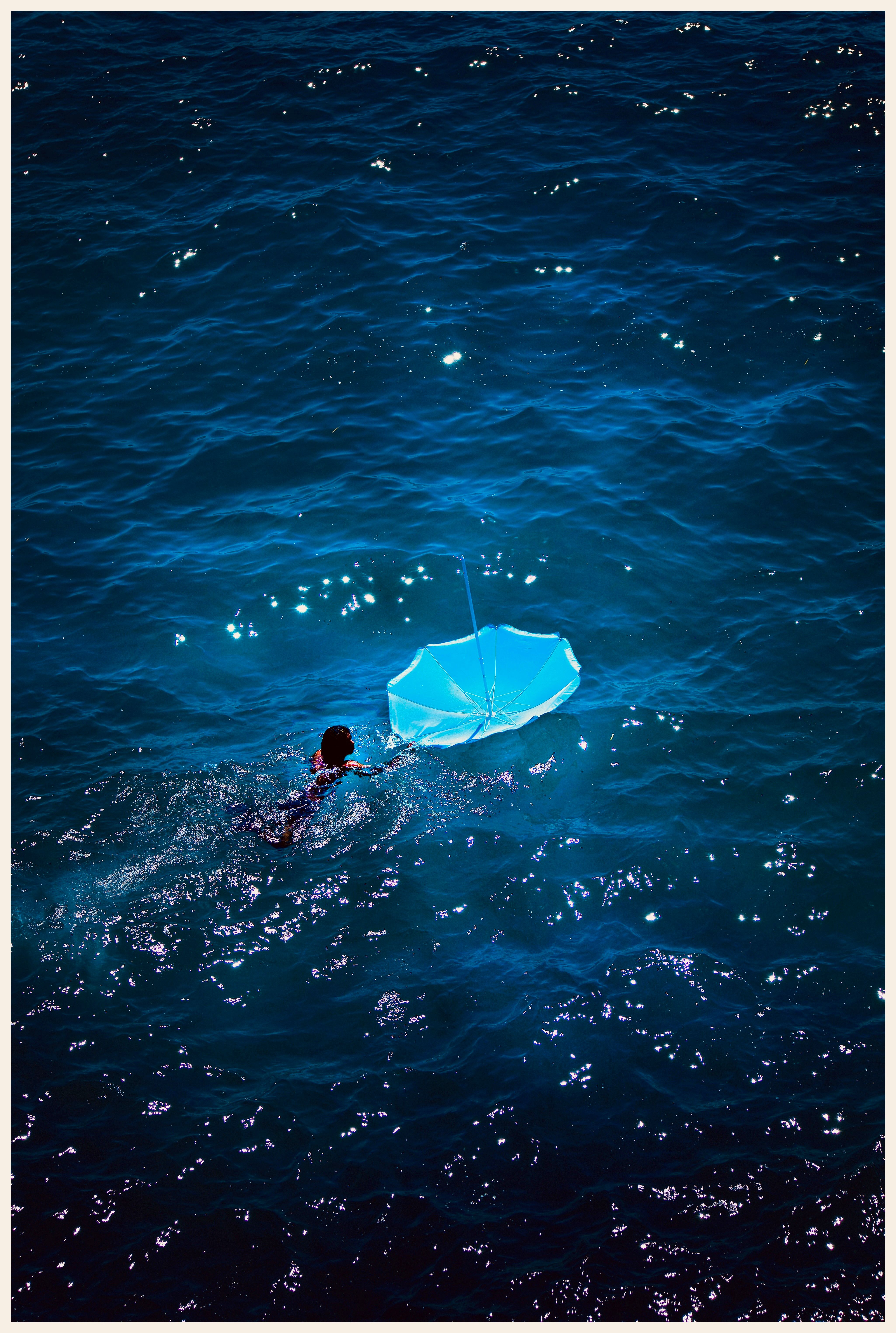 Un nageur tire un parasol bleu retourné sur une mer sombre et scintillante